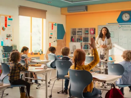 Teacher explaining a math lesson to classroom full of children and one girl is raising her hand